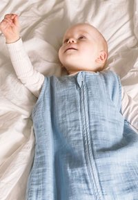 Light blue, sleeveless baby sleep sack with a front zipper and textured fabric, laid on a white surface, with a child reaching up.