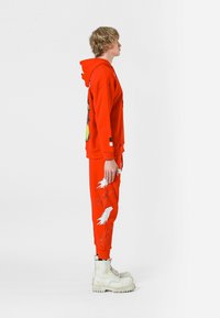 Sudadera con capucha roja con diseño gráfico, combinada con pantalones de chándal rojos que presentan patrones de plumas blancas, llevados con botas blancas gruesas.