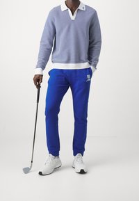 Sininen ja valkoinen raidallinen pitkähihaisena polo-paita, V-kaula-aukolla, yhdistettynä sinisiin housuihin ja valkoisiin urheilusandaaleihin, pitäen golf-mailaa.