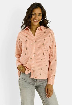Mujer que lleva una camisa de botones a rayas rosa y blanca con pequeños diseños bordados y vaqueros gris claro, sonriendo con una mano en el bolsillo.