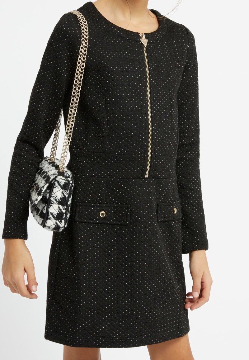 Femme portant une robe noire à manches longues avec une fermeture éclair dorée et de petits points, tenant un sac à main noir et blanc à motifs avec des anses en chaîne dorée.