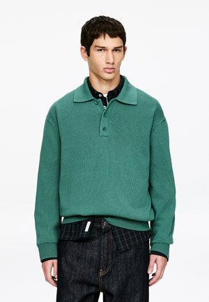 Młody mężczyzna ubrany w zielony dziergany sweter na czarnej koszuli w kratę i ciemnych dżinsach, stojący na tle jednolitego białego tła.