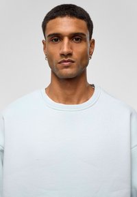  Lichtblauwe sweatshirt met een ronde hals en verlaagde schouders. Gladde textuur, minimalistisch ontwerp, gedragen over een wit T-shirt.