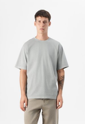 Un mannequin masculin se tient contre un fond blanc, portant un t-shirt oversize gris clair et un pantalon beige, avec un tatouage visible sur son bras.