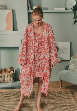 Frau, die lächelnd nach unten schaut und ein lockeres, langes, florales pink-weißes Kleid trägt, barfuß in einem Wohnzimmer steht.