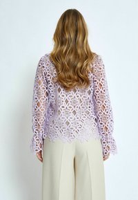 Top in pizzo lilla con motivo geometrico aperto, maniche lunghe, orlo scallop, abbinato a pantaloni larghi crema. Testurizzato e trasparente.