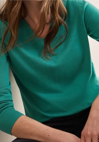 Turquoise long-sleeve knit sweater with a V-neck, featuring ribbed cuffs and a smooth texture. Paired with black pants.