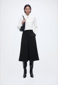 Γυναίκα με λευκό πουκάμισο με φιόγκο, μαύρη μίντι φούστα, μαύρες μπότες μέχρι το γόνατο και κρατώντας μια μαύρη τσάντα ώμου.