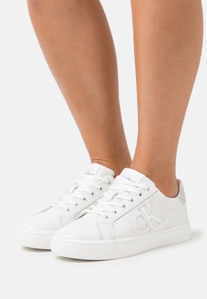 Jambes portant des baskets basses blanches à lacets, logo CK sur le côté et texte « Calvin Klein Jeans » sur la semelle et la languette.