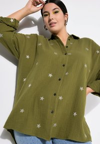 Olijfgroene button-up shirt gemaakt van textuurstof met zilveren sterrenborduring. Beschikt over een losse pasvorm en zwarte knopen. Korte, opgerolde mouwen.