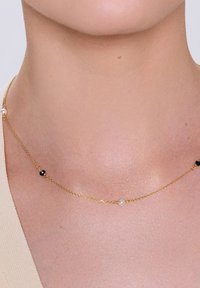 Złoty łańcuszek z naprzemiennie rozmieszczonymi białymi perłami i małymi czarnymi koralikami, delikatny i cienki w designie, spoczywający na szyi noszącej go osoby.