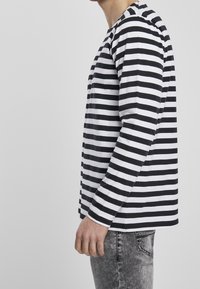 Camicia a maniche lunghe a righe bianche e nere in cotone, con scollo a giro e taglio dritto, abbinata a jeans grigi strappati.