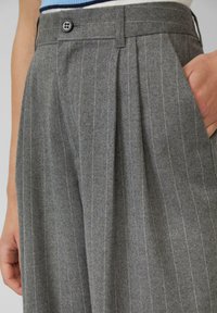 Pantaloni grigi a righe con pieghe, cintura con bottoni, tasche laterali, indossati da una persona con una mano in tasca e orlo di una camicia a righe visibile.