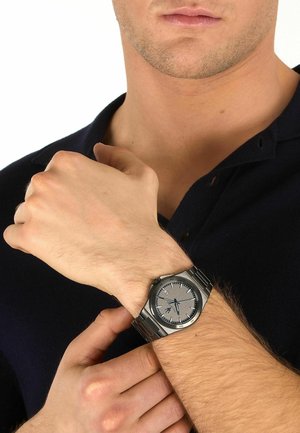 Uomo con camicia nera che regola un orologio da polso in metallo argento con quadrante grigio sul polso sinistro.