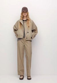 Beige kort jacka med stora knappar över en randig topp, tillsammans med vida beige byxor och bruna sneakers, avslutad med en brun keps.