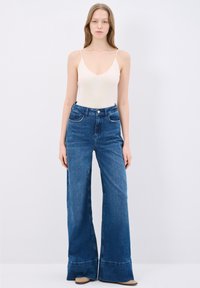 Bodysuit de color claro con tirantes finos, combinado con unos vaqueros oscuros de tiro alto y pernera ancha, que presentan bolsillos frontales y un dobladillo deshilachado.