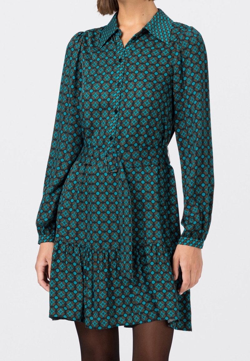 Femme portant une robe à manches longues à motif bleu sarcelle et noir, avec un col et un devant boutonné, debout devant un fond clair uni.