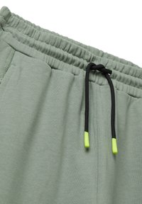 Hellgrüner Sweatpants-Bund mit gerafftem Elastik, schwarzer Zugkordel mit neongelben Enden und sichtbarem Stich der Seitentasche.