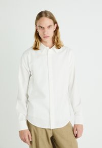 Chemise blanche à manches longues avec un col classique, fabriquée en coton, et ornée d'une broderie subtile sur la poitrine gauche. Texture lisse.