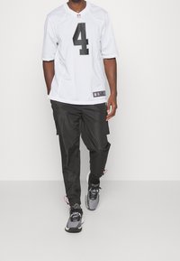 Hombre caminando con una camiseta blanca de la NFL con el número 4, pantalones cargo negros y zapatillas deportivas grises y negras contra un fondo blanco.