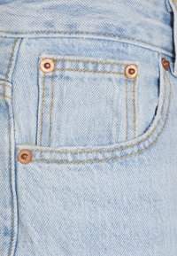 Um tecido de denim azul claro apresenta um pequeno bolso, costura visível e rebites de cobre. A textura é suave com um leve desbotamento.
