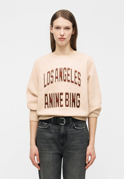 Femme aux cheveux bruns raides portant un sweat-shirt beige avec le texte "LOS ANGELES ANINE BING" et un jean noir, debout devant un fond blanc.