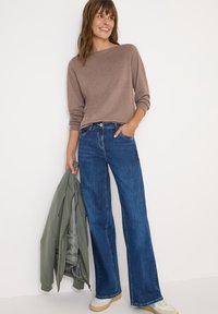 Maglietta marrone a maniche lunghe abbinata a jeans svasati blu scuro. La modella tiene in mano un bomber verde e indossa sneakers beige.