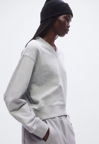 Harmaa puuvillainen collegepaita, jossa on V-aukko, ylivertainen istuvuus ja pitkät hihat, yhdistetty samansävyisiin collegehousuihin. Päällä musta neulottu pipo.