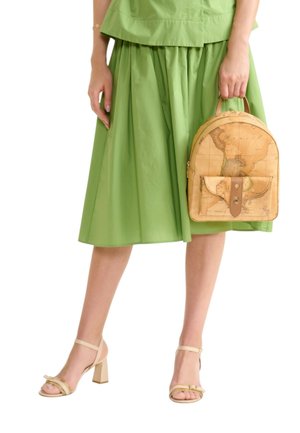 Donna con gonna verde e sandali beige con tacco, che tiene uno zainetto piccolo color cuoio con un design di mappa d'epoca.