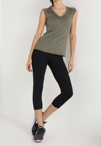 Oliwkowy top bez rękawów z dekoltem w serek, zestawiony z dopasowanymi czarnymi leginsami Capri. Model nosi czarno-szare buty sportowe.