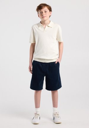 Jongen met lichtbruin haar die een crèmekleurig poloshirt met korte mouwen draagt, donkerblauwe korte broek, witte sokken en witte sneakers, staand met één hand in de zak.