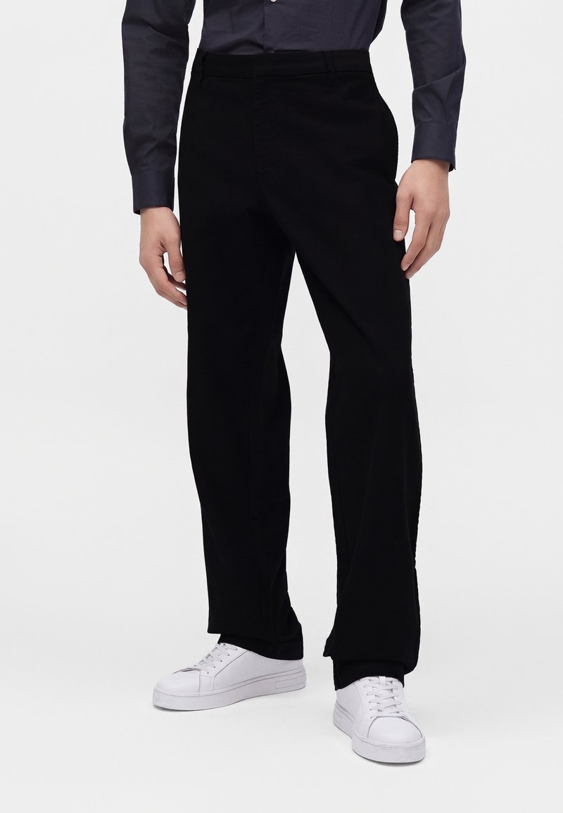 Homme portant un pantalon noir droit, une chemise sombre à manches longues et des baskets blanches, debout devant un fond uni.