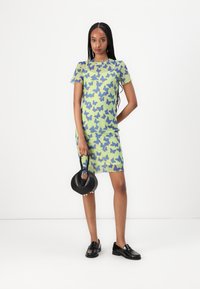 Robe à manches courtes de couleur vert clair avec un motif de papillon bleu et orange, associée à des chaussures noires à enfiler et un petit sac à main noir.