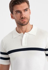 Vitt stickat polo-shirt med en treknappslå och marinblå horisontella ränder på ärmarna. Slät textur, klassisk passform.