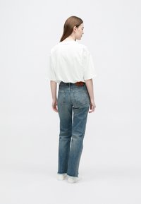 Mujer con camiseta blanca de gran tamaño, vaqueros azules de pierna recta con dobladillos deshilachados y zapatillas blancas, de pie de espaldas sobre un fondo blanco liso.