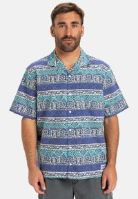 Camisa de botones de manga corta en azul y púrpura con intrincados patrones geométricos y florales, hecha de una tela ligera con un corte relajado.