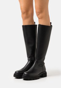 Black knee-high boots made of smooth leather, featuring a side zipper, thick rubber sole, and rounded toe design. Easy pull-on tabs.