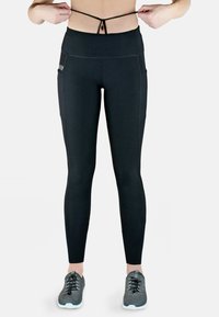 Schwarze high-waist Leggings mit glatter Textur, ausgestattet mit Seitentaschen und einem Kordelzugdetail an der Taille. Getragen mit grauen Turnschuhen.
