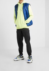 Niebieska kamizelka puchowa, neonowo-zielona bluza z kapturem, czarne spodnie cargo oraz wielokolorowe buty sportowe z teksturowanymi detalami. Casualowy, warstwowy strój.