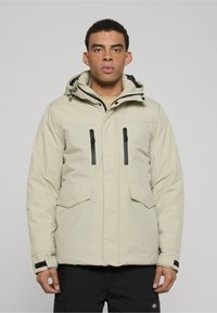 Beige insulated jacket with a hood, featuring two zippered chest pockets and adjustable cuffs for enhanced fit and functionality.