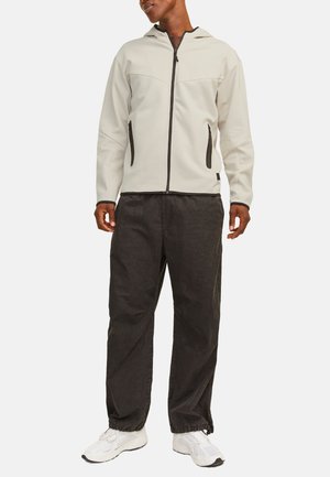 Grijze rits-hoodie met een hoge kraag, zwarte accenten en zijzakken, in combinatie met losse zwarte broeken en witte sneakers.