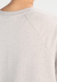 Tissu en jersey gris clair, doux, avec un col rond et des manches raglan. Texture lisse, avec des variations subtiles de teinte.