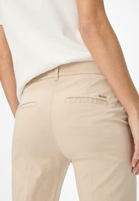 Pantalon beige en mélange de coton avec une coupe droite, doté de deux poches arrière et d'un petit accent logo métallique. Texture lisse.