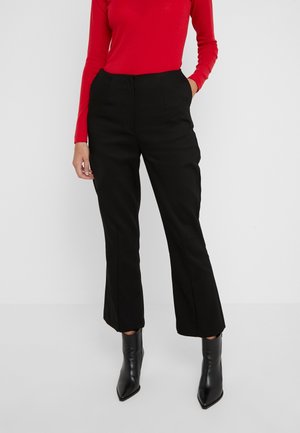 Osoba ubrana w czarne, wysokim stanem i prostymi nogawkami spodnie, czerwony top z długim rękawem oraz czarne, szpiczaste botki do kostki, stojąca z jedną ręką w kieszeni.