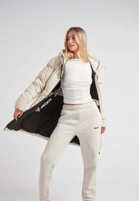Chaqueta tipo plumas en color beige claro, con un interior negro que presenta un logo. Combinada con una camiseta blanca ajustada y pantalones de chándal beige claro con marca.