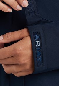 Veste marine en tissu texturé avec une finition lisse ; comprend une étiquette tissée avec "Ariat" en bleu. Gros plan de la main et de la zone du poignet.