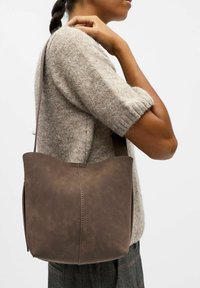 Personne portant un pull beige texturé et un pantalon à carreaux, portant un grand sac en cuir marron à bandoulière avec un bord supérieur arrondi.