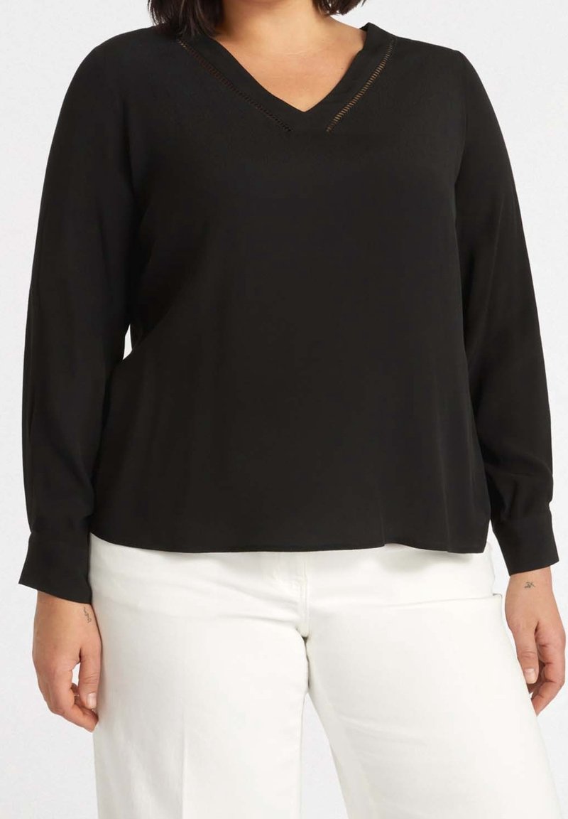 Femme portant un haut noir à manches longues à col en V, associé à un pantalon blanc, affichant un style de mode décontracté et minimaliste.