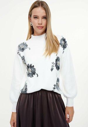 White sweater with floral sequin embellishments in silver and black; features a high neckline and puffed sleeves, paired with a dark skirt.