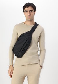 Bolsa de bandolera negra y ligera con cremalleras diagonales y una sola correa para el hombro, llevada sobre una blusa de manga larga beige y pantalones a juego.